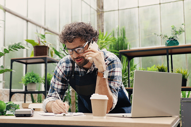 Happy Caucasian businessman talking on cellphone while standing in apron in small floral center and writing down order details. Joyful male florist calling on smartphone at work.